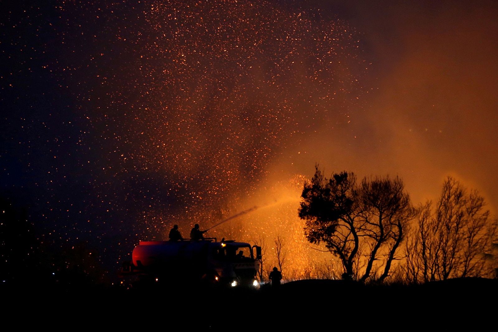 Ein Feuerwehrauto löscht nachts einen Brand im Wald. Vor dem Funkenregen steht ein Baum