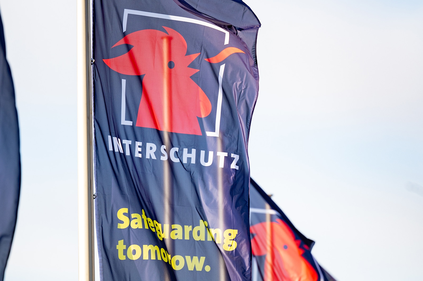 Eine INTERSCHUTZ Fahne weht an einem Mast. Neben dem Logo ist der Claim zu sehen: Safeguarding tomorrow