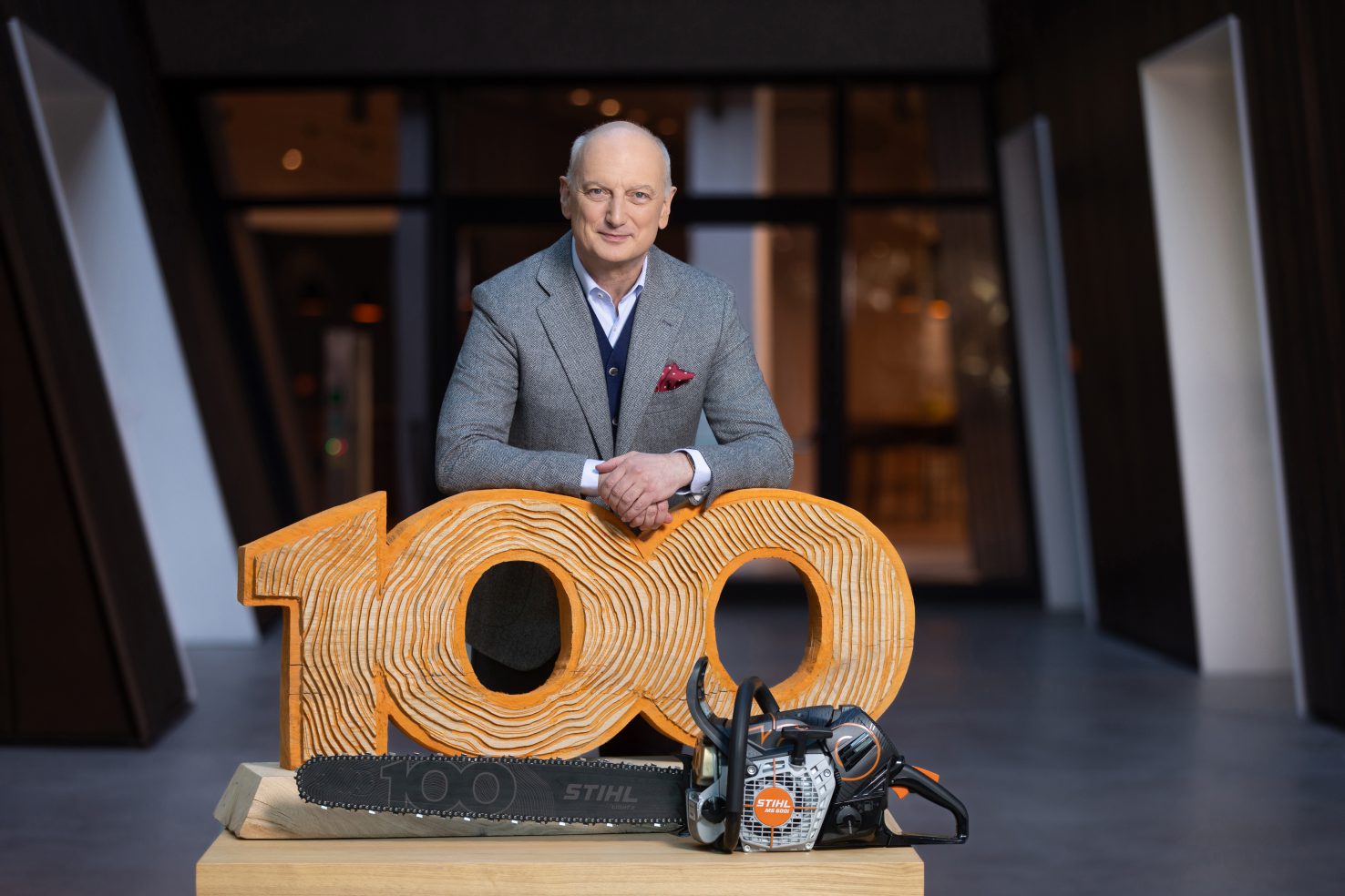A man in a suit leans on a wooden sculpture shaped like the number “100,” with a chainsaw placed in front—likely a staged anniversary display.
