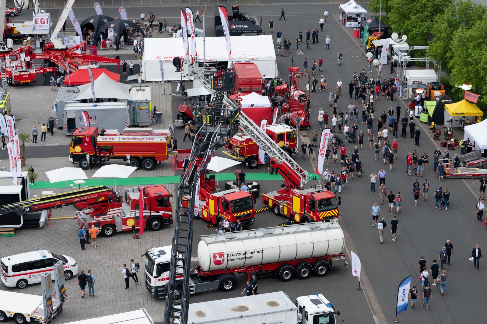 Aerial view with many fire and emergency vehicles, extended ladders, and booths; numerous visitors are walking between the exhibition areas.